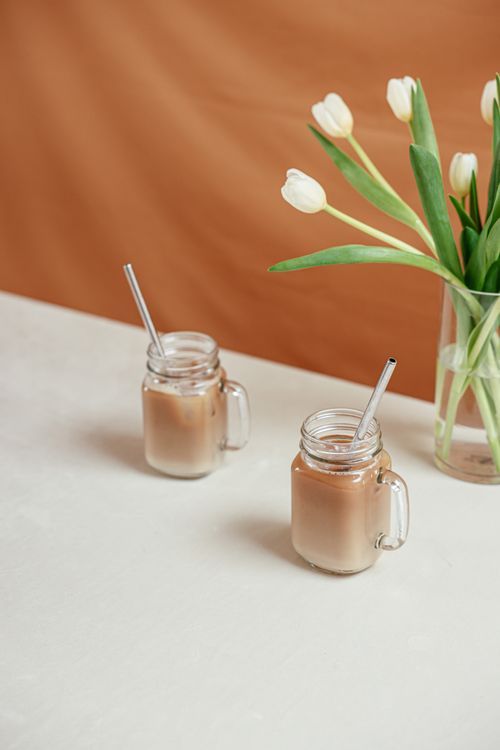Free Two iced coffee beverages in mason jars with metal straws and white tulips in a vase on a table. Stock Photo