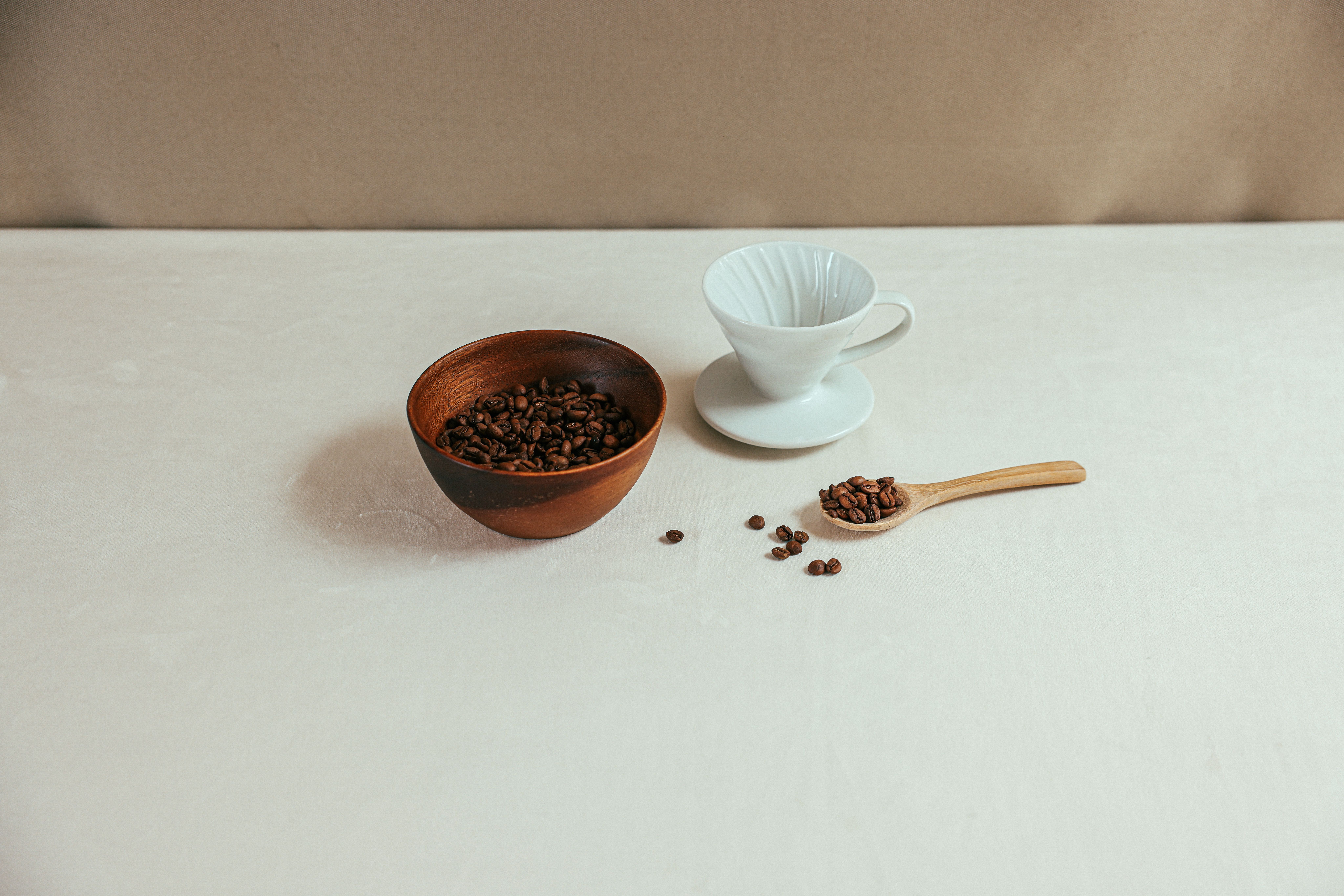 A simple coffee setup featuring a ceramic cup, wooden bowl, and coffee beans on a light table.