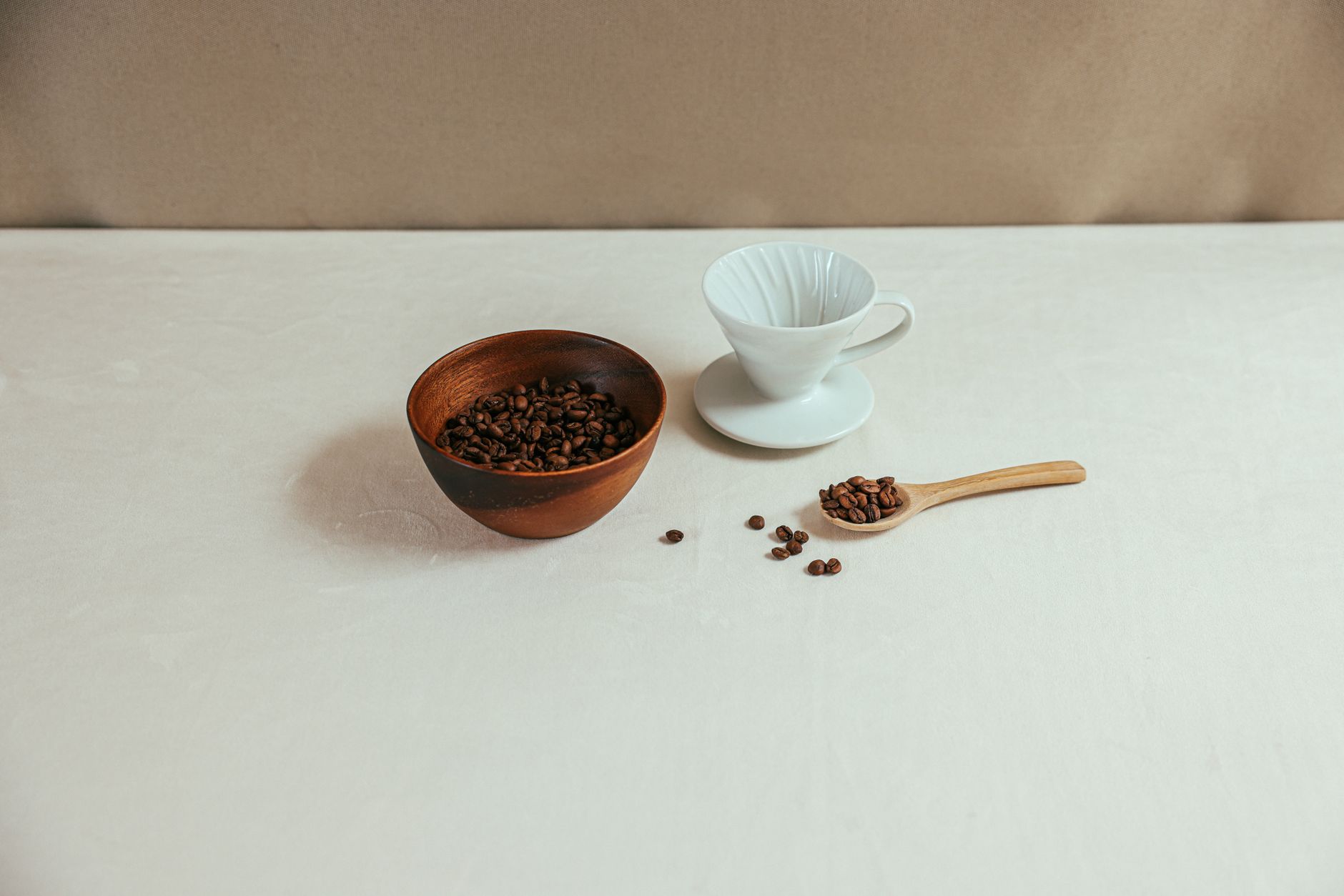 A simple coffee setup featuring a ceramic cup, wooden bowl, and coffee beans on a light table.