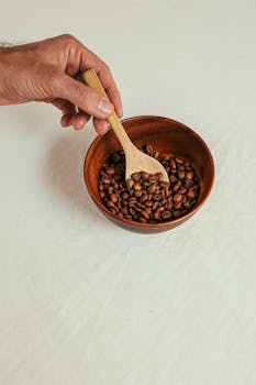 A hand holding a wooden spoon with aromatic coffee beans in a wooden bowl on a white table.
