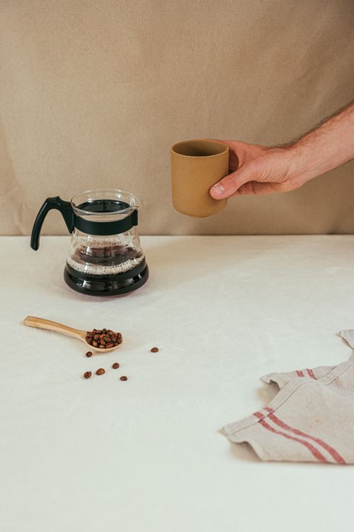 Free A minimalist coffee setup with a hand holding a mug, coffee pot, beans, and napkin on a white surface. Stock Photo