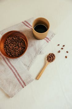 Flat lay of coffee beans in bowl and cup, set on a striped towel with wooden spoon.