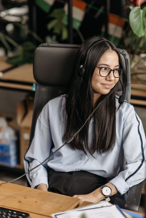 Free Asian woman in blue shirt wearing headphones in an office setting, smiling. Stock Photo