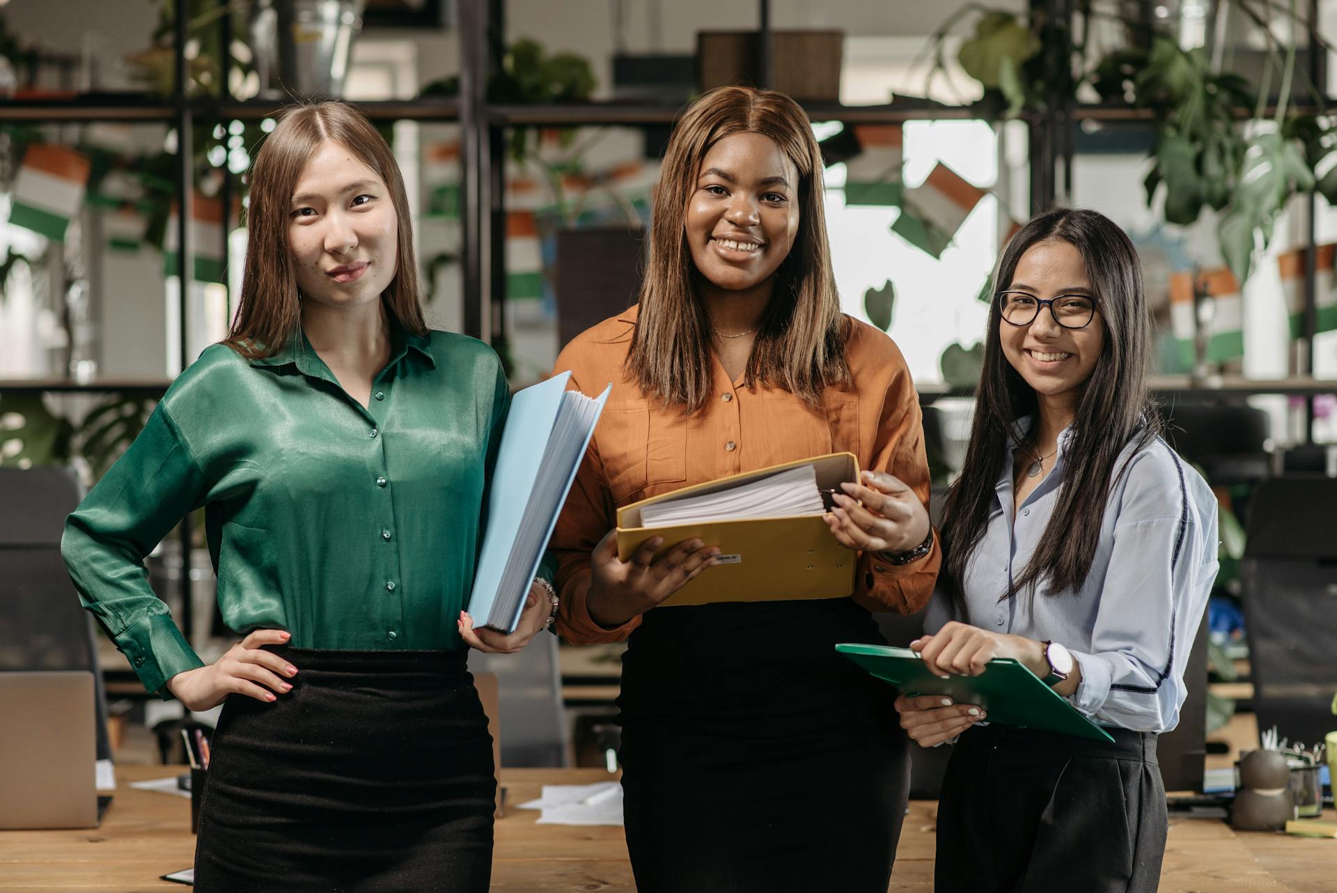 Three diverse businesswomen standing together in an office, holding files and smiling.