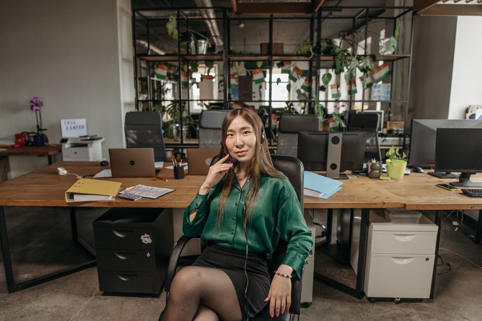 Asian woman in green blouse and headphones sits at modern office desk, call center environment.