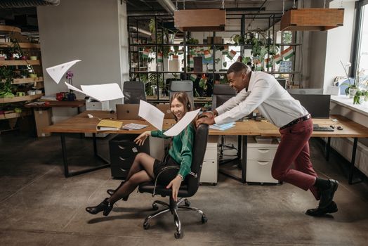 Two colleagues having fun in the office, pushing a chair amidst a lively work environment.