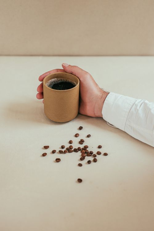 Free A person's hand holding a rustic cup of black coffee with scattered coffee beans on a white surface. Stock Photo