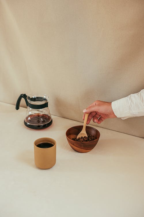Woman Holding a Spoon in a Bowl with Coffee Beans next to a Coffee Pot and a Coffee Cup 