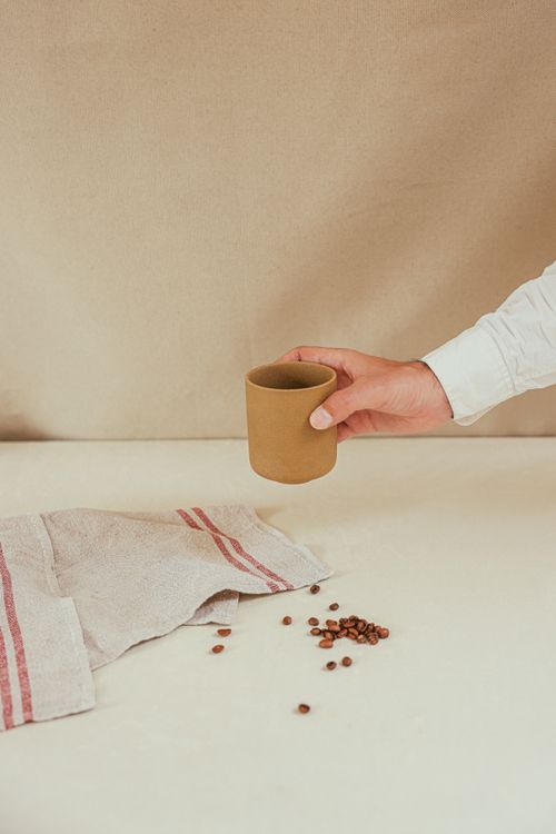 Free A minimalist image featuring a hand holding a coffee cup beside scattered beans on a cloth. Stock Photo