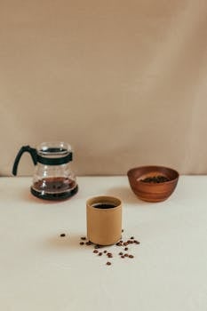 Coffee cup with beans, glass pot, and wooden bowl on a beige surface.
