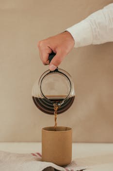 A close-up image of a hand pouring coffee from a glass pitcher into a cup on a neutral background.