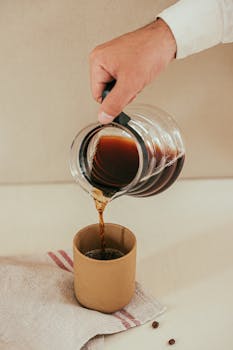 A hand pours aromatic filter coffee from a pot into a cup on a white surface.