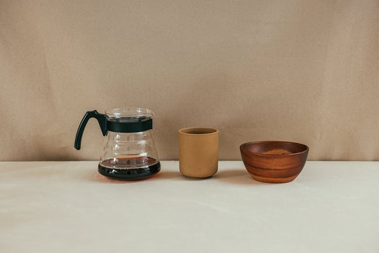 A Brown Cup Between A Clear Glass Pitcher With Coffee And A Wooden Bowl