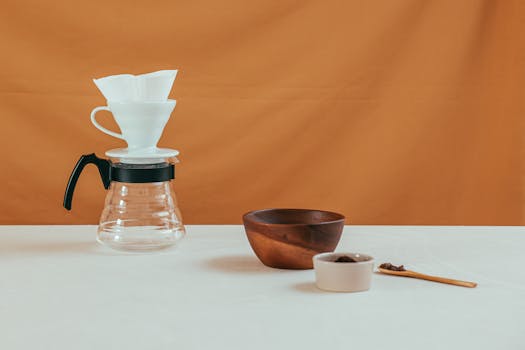 Minimalist coffee setup with pour over, bowls, and spoon on white table against orange background.