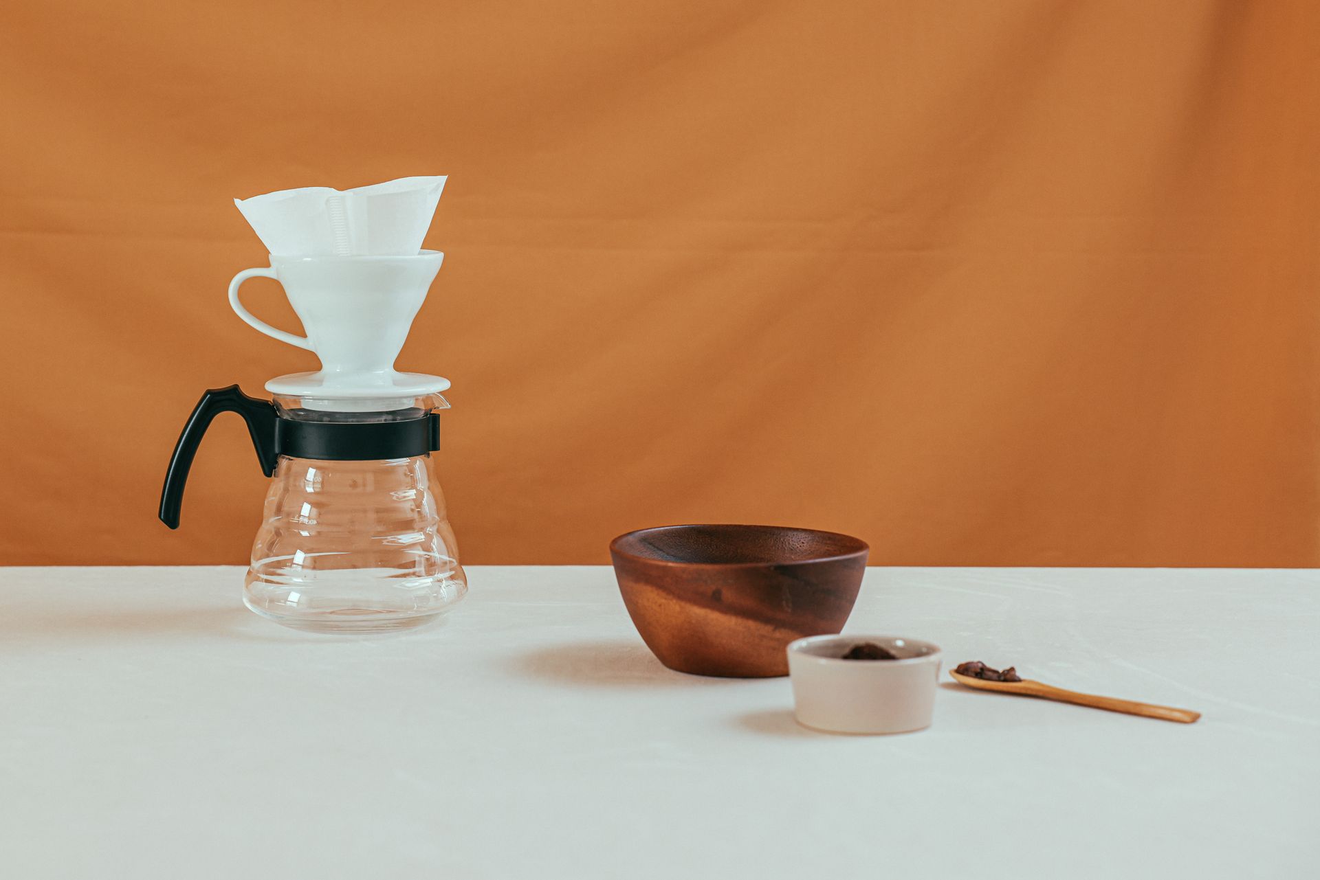 Minimalist coffee setup with pour over, bowls, and spoon on white table against orange background.
