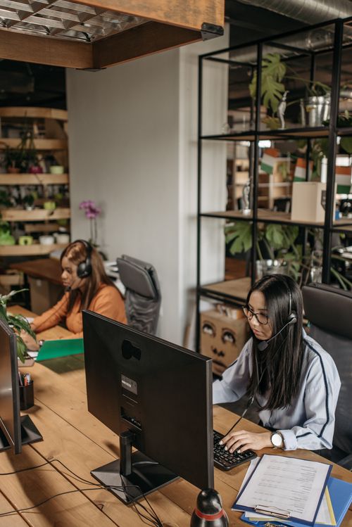 Free Two women in headsets working diligently in a modern office space with green plants and industrial design elements. Stock Photo
