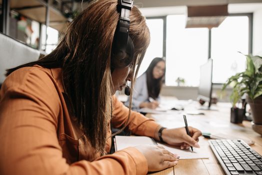 Side view of a dedicated businesswoman with headphones working at her desk, taking notes.