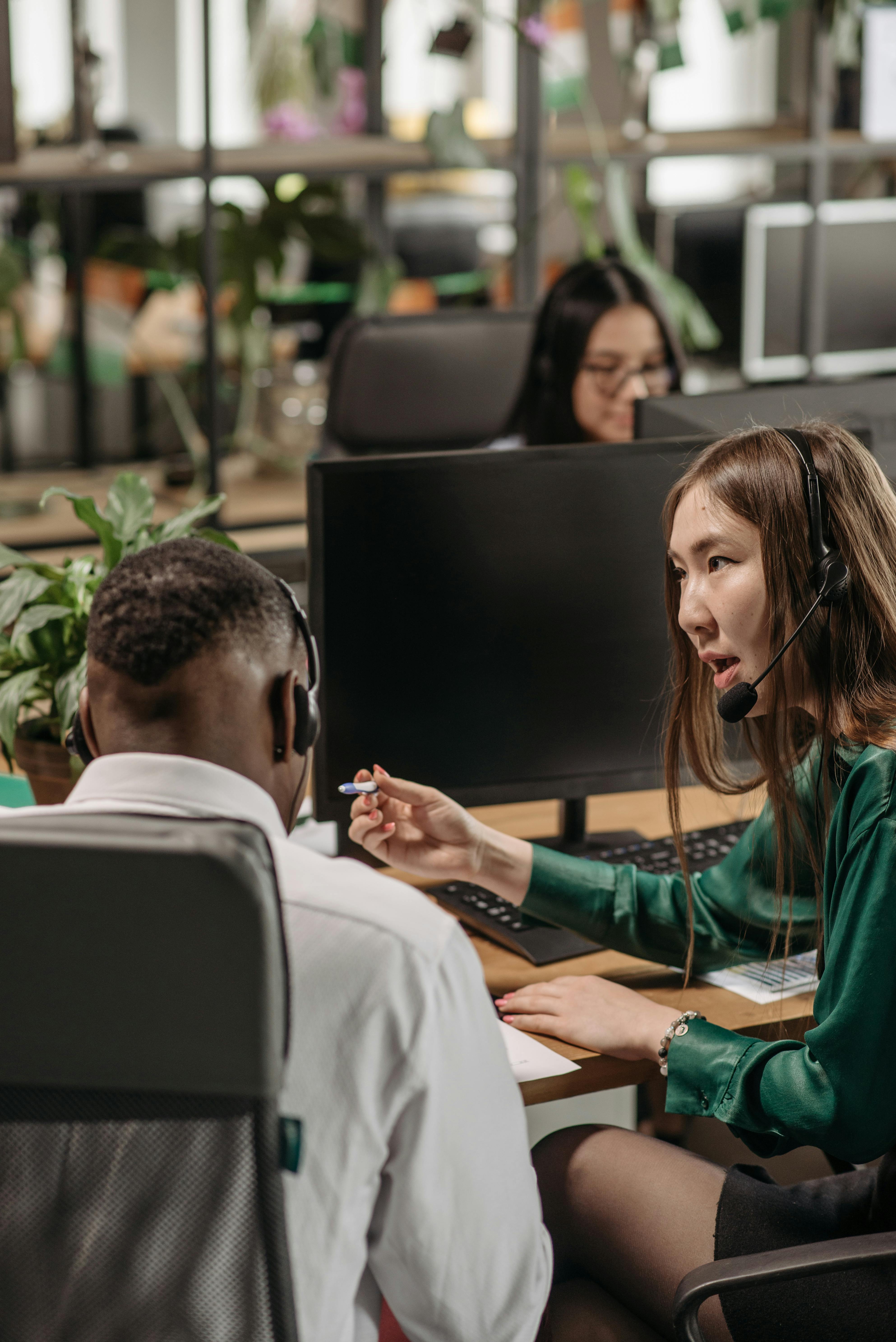 Colleagues wearing headsets discuss work at computer desks in a modern office.