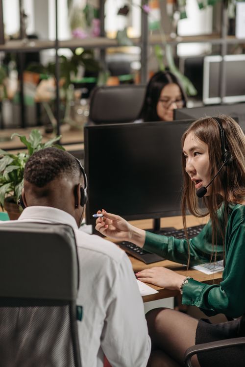 Free Colleagues wearing headsets discuss work at computer desks in a modern office. Stock Photo