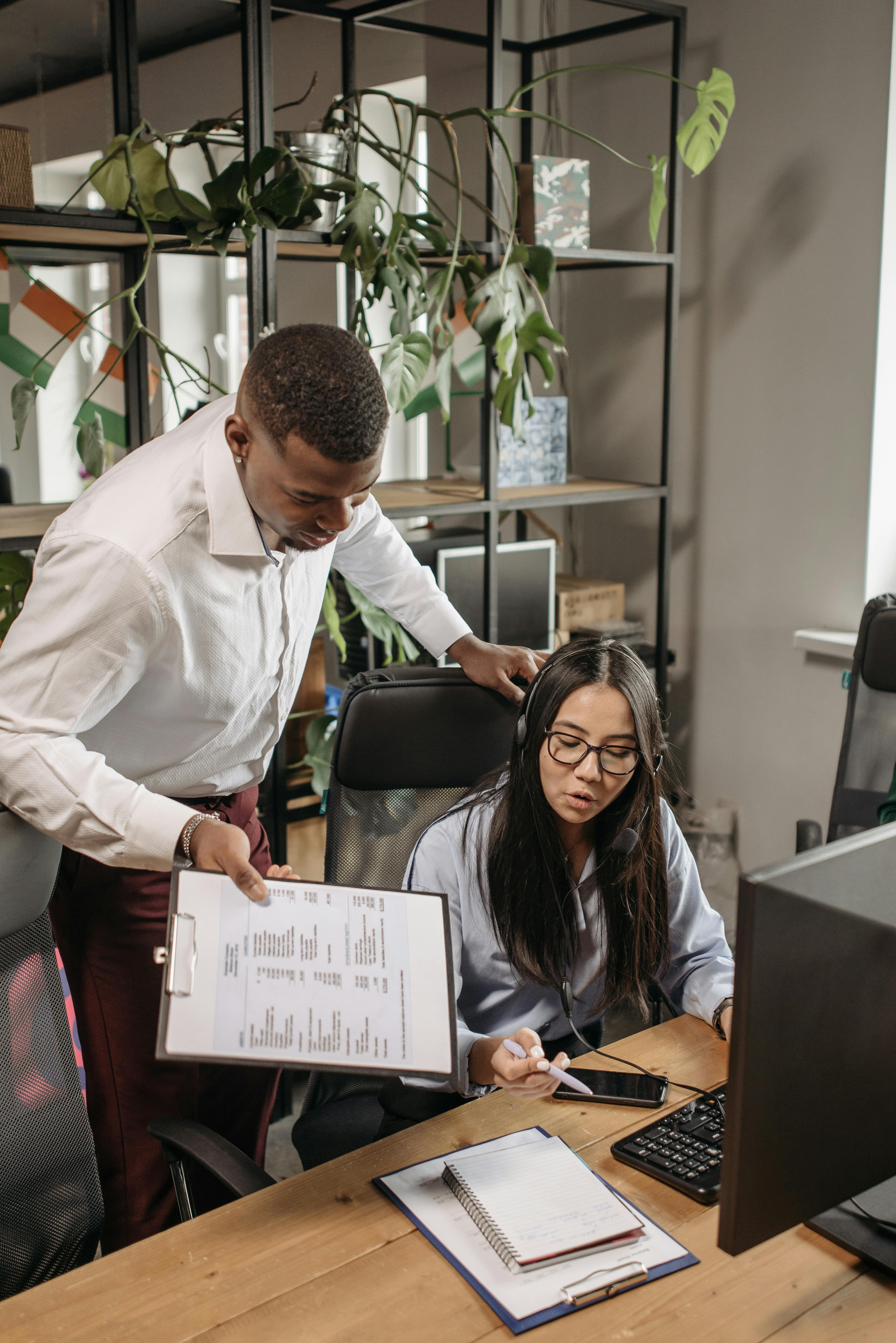 Two office workers collaborate over documents and a computer in a modern workspace.