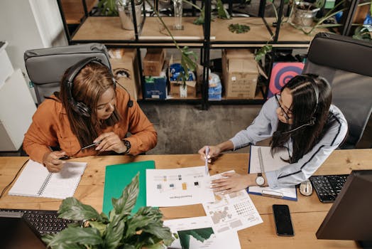 Two women wearing headsets discuss work at a shared desk in an office.