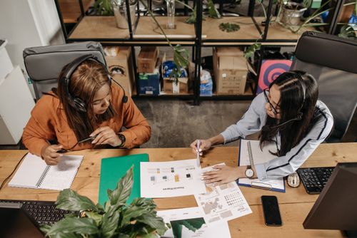Free Two women wearing headsets discuss work at a shared desk in an office. Stock Photo