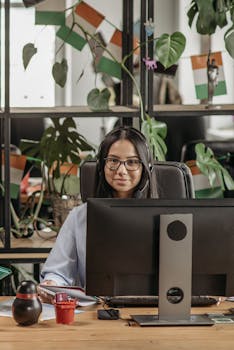 Asian woman with eyeglasses working at a desk in a modern office, surrounded by plants.