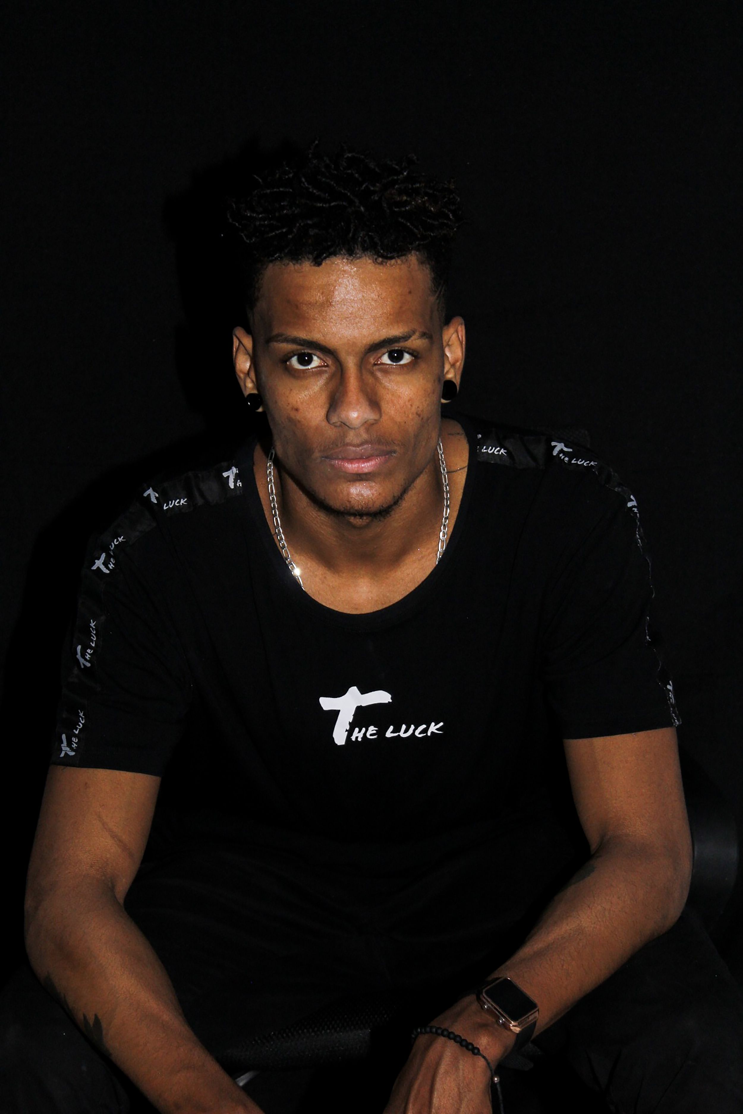 Homem confiante com camiseta preta posando contra um fundo escuro.