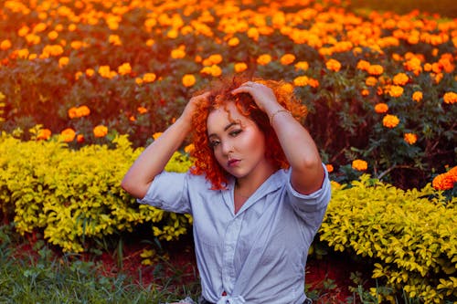 Free Trendy female with makeup in shirt touching curly hair while looking at camera against colorful blossoming flowers in garden Stock Photo