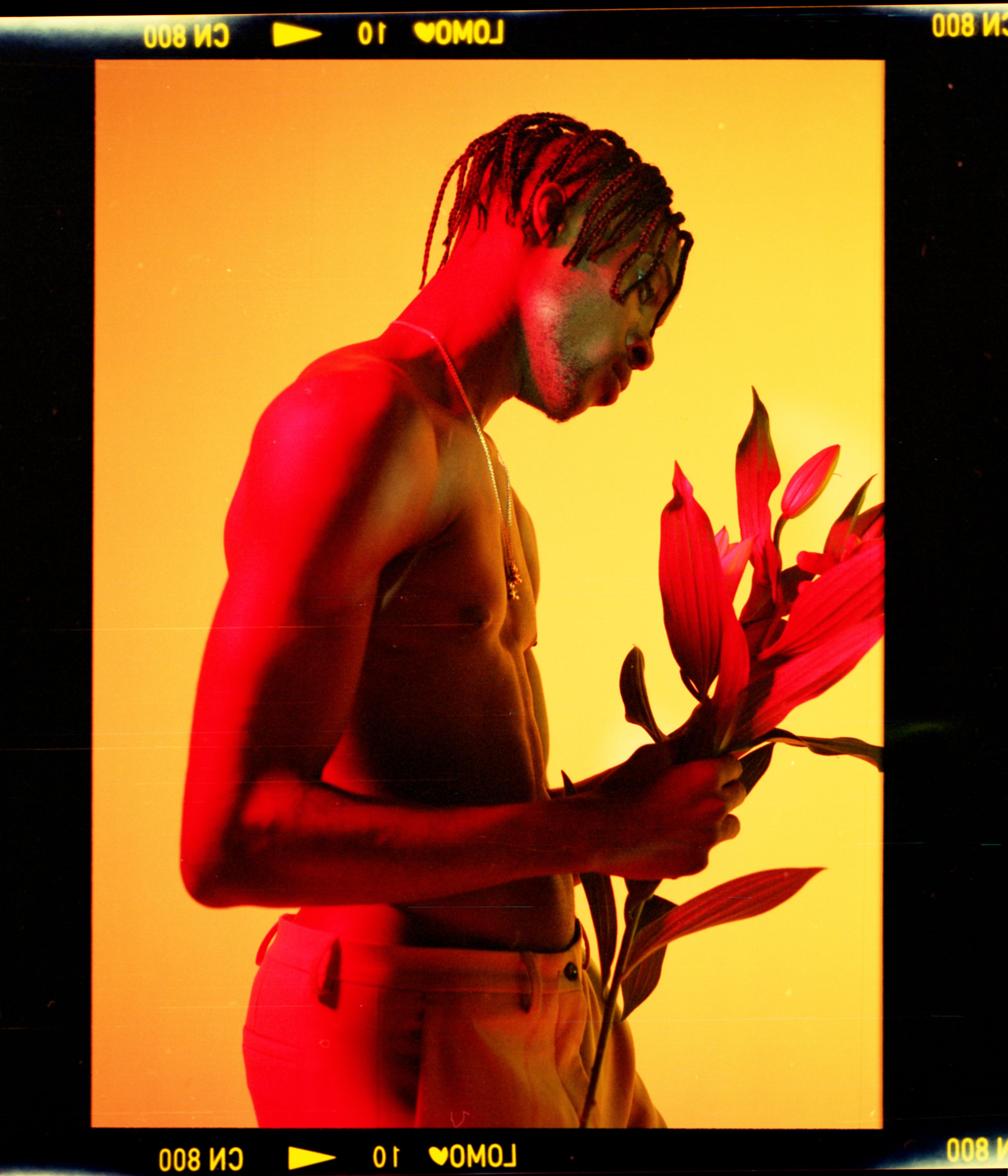 Artistic portrait of a topless man with braided hair holding flowers against a vibrant yellow background.