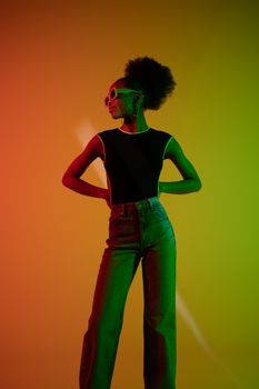 Fashionable woman with curly hair and sunglasses poses confidently against vibrant background.