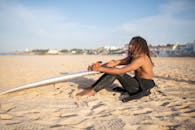 一个光着膀子的男性冲浪者坐在沙滩上，在明媚的阳光下拿着冲浪板