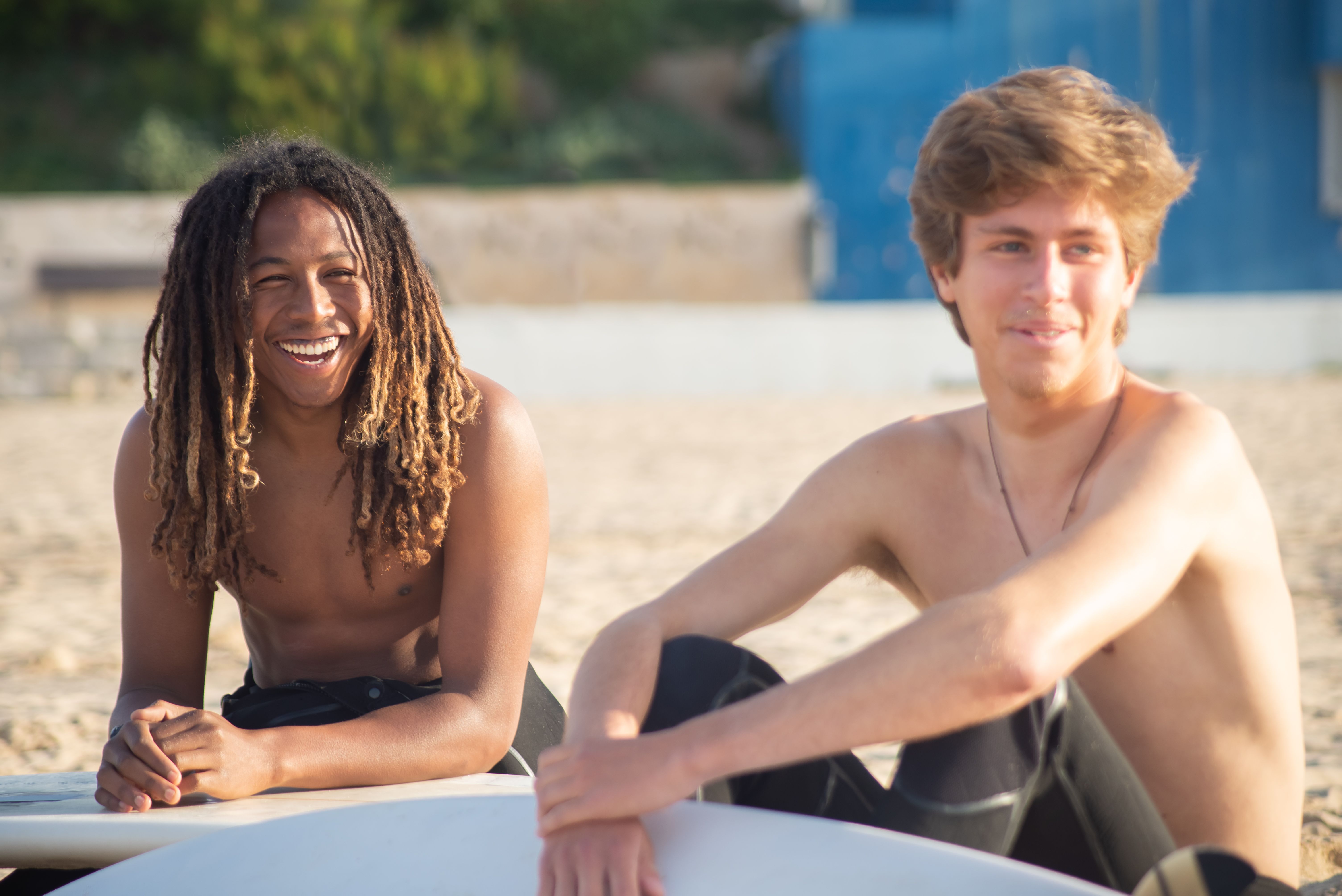 Two young men sitting on the beach with surfboards, enjoying a sunny day and smiling.