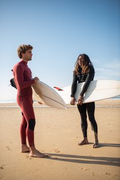 Two surfers in wetsuits with boards on a sunny Portugal beach.
