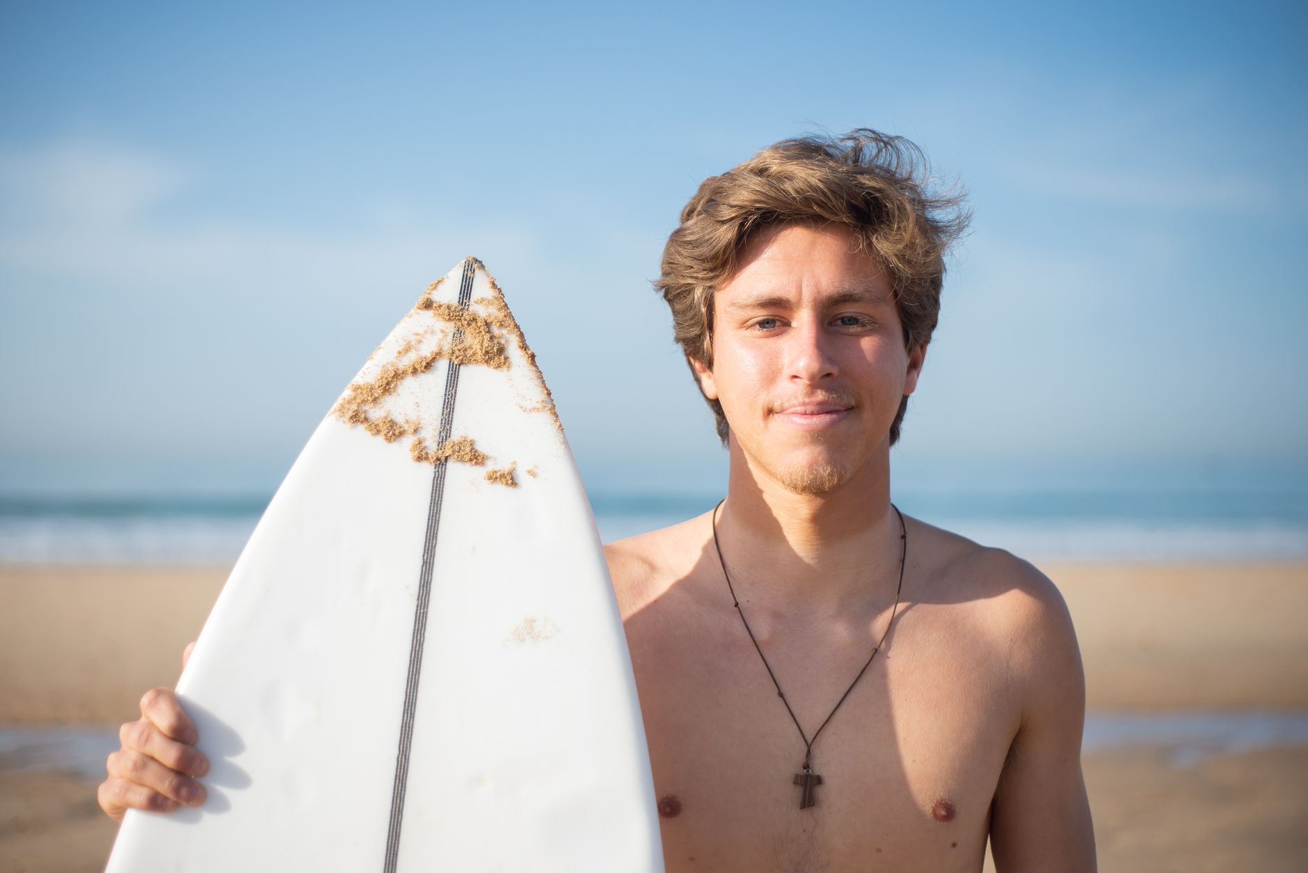 Young shirtless man holding a surfboard on a sunny beach in Portugal, summer vibes.