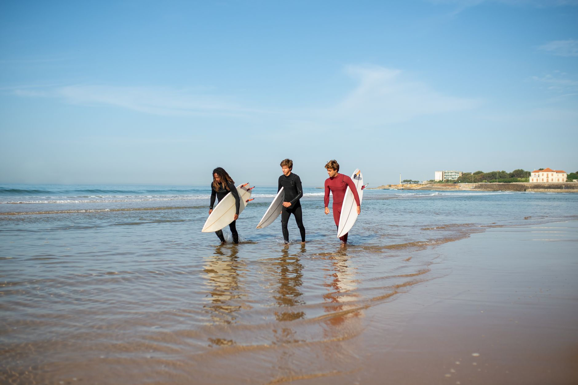 Three surfers in wetsuits carry surfboards on a sunny beach in Portugal.