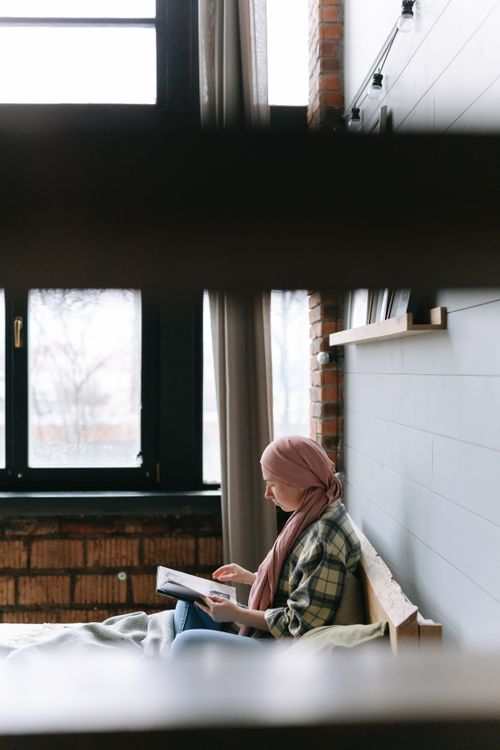 Free A woman in a head wrap reads a book by a window in a cozy, well-lit bedroom. Stock Photo