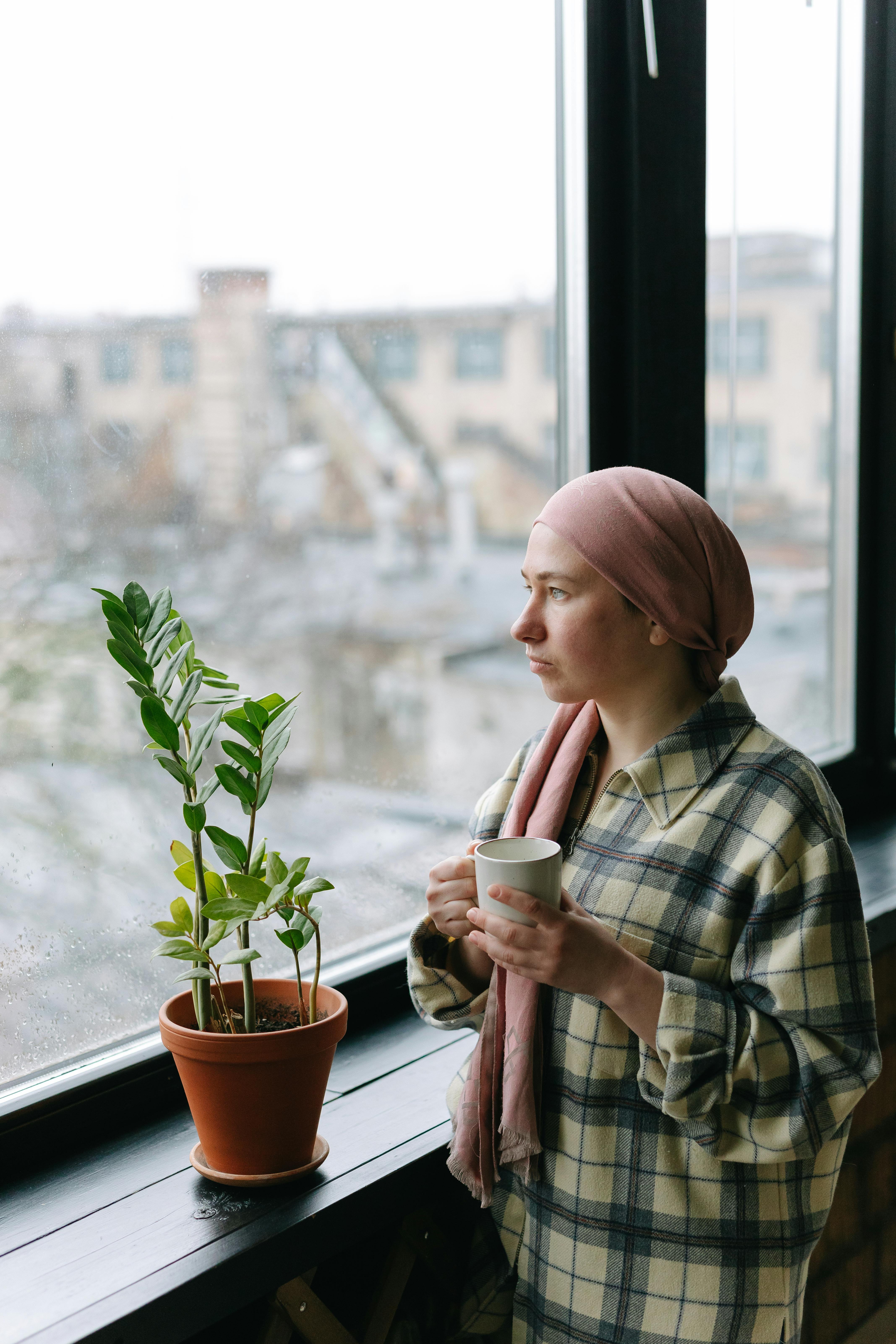 Woman in head cover holding coffee by a window, indoors with a potted plant.