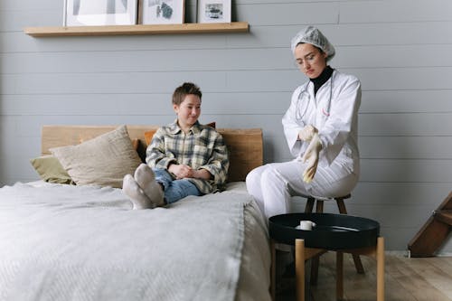 Patient sits on bed interacting with a doctor in a cozy bedroom environment.