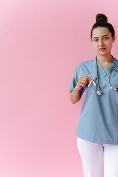 Medical professional holding pink ribbon for breast cancer awareness against light pink background.