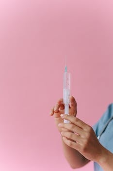 Close-up of hands holding a syringe against a pink backdrop, medical theme.