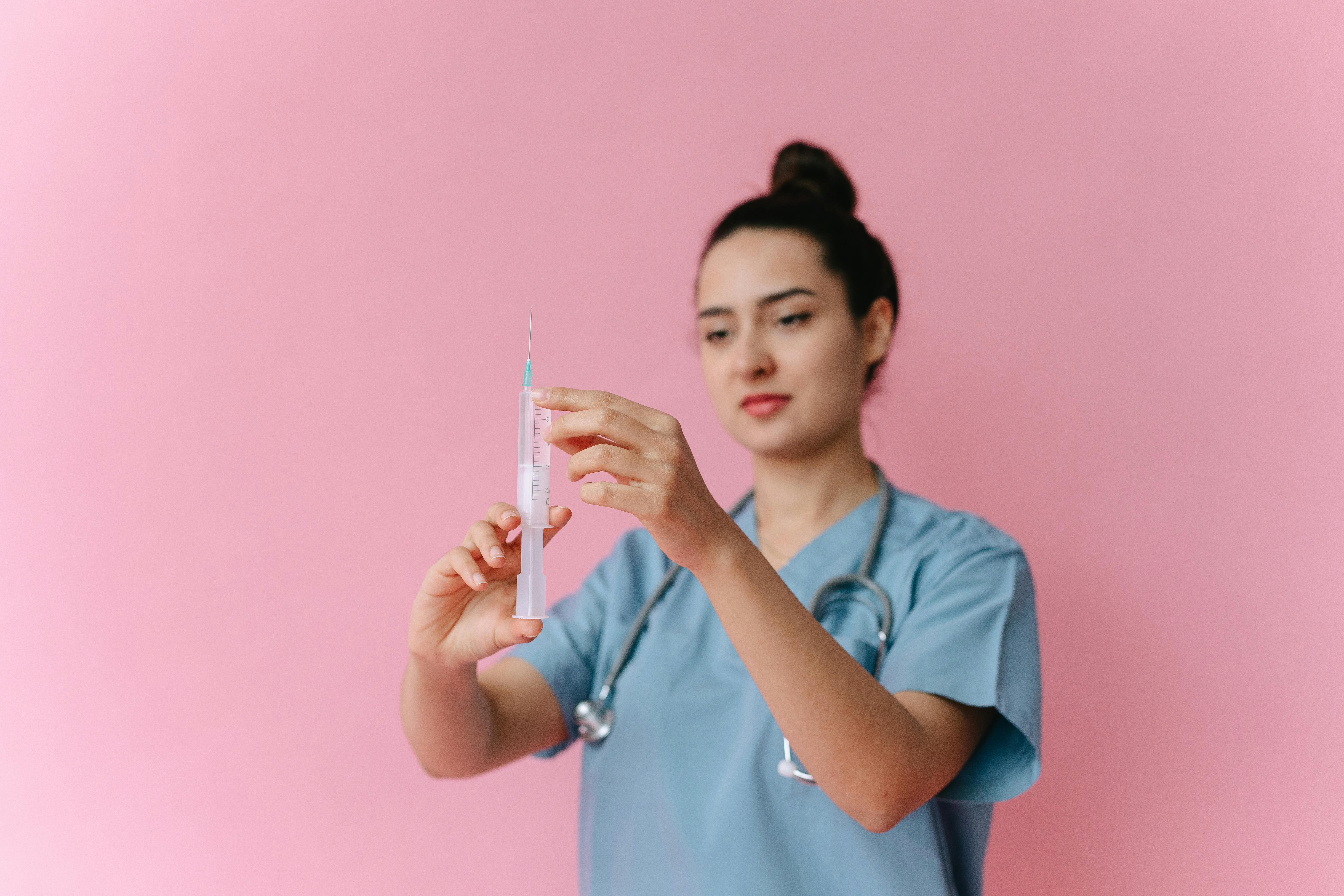 Female medical practitioner holding a syringe, standing in front of a pink background.