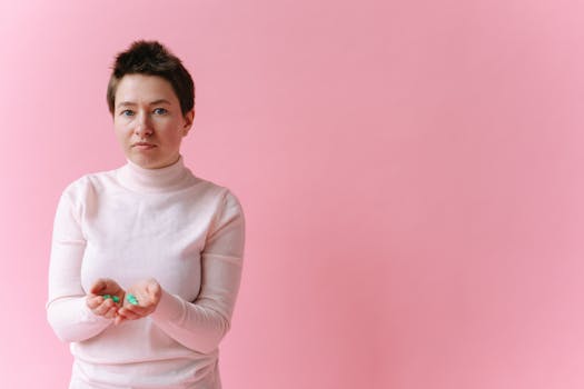 A Caucasian woman holds pills in her hands against a soft pink backdrop, conveying healthcare themes.