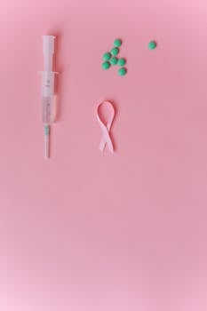 Flat lay of a syringe, pink ribbon, and green pills on a soft pink background symbolizing breast cancer awareness.