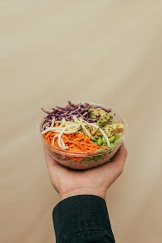 Close-up of a hand holding a vibrant rice bowl with vegetables on a beige background.