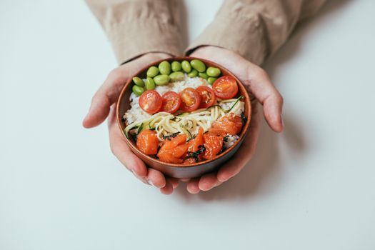 Close-up of hands holding a salmon poke bowl with cherry tomatoes, edamame, and rice.