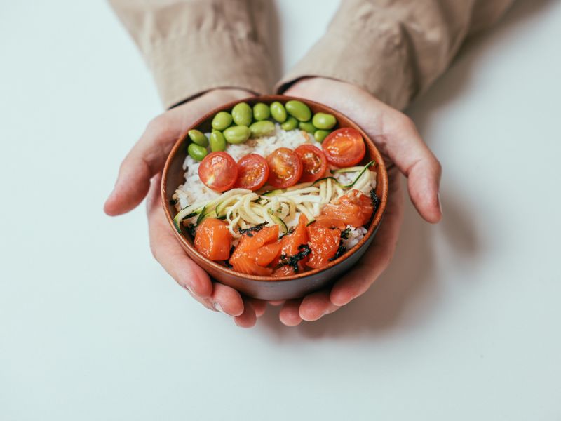 Close-up of hands holding a salmon poke bowl with cherry tomatoes, edamame, and rice.