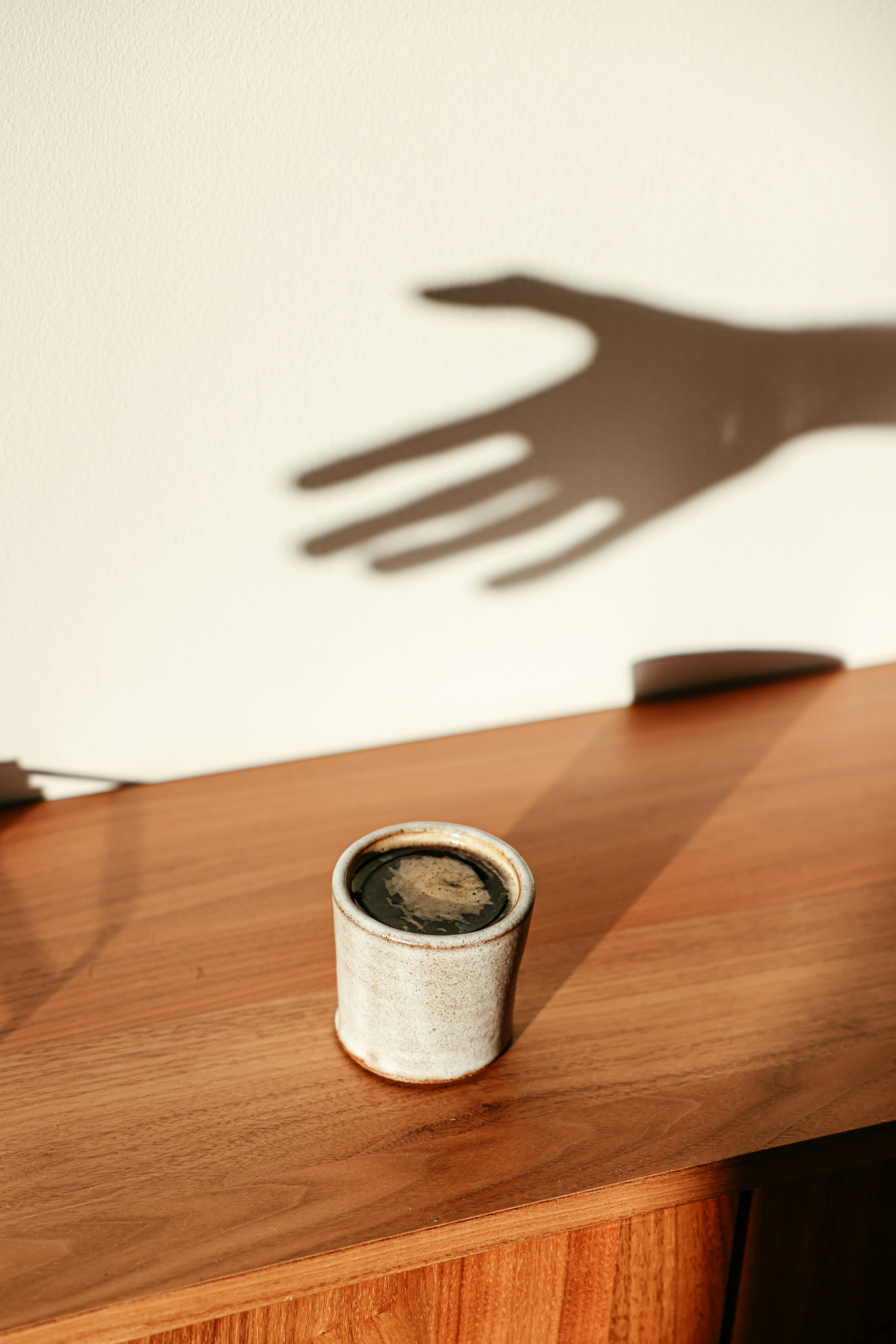 A coffee cup on a wooden table with an artistic shadow play of a hand in soft lighting.