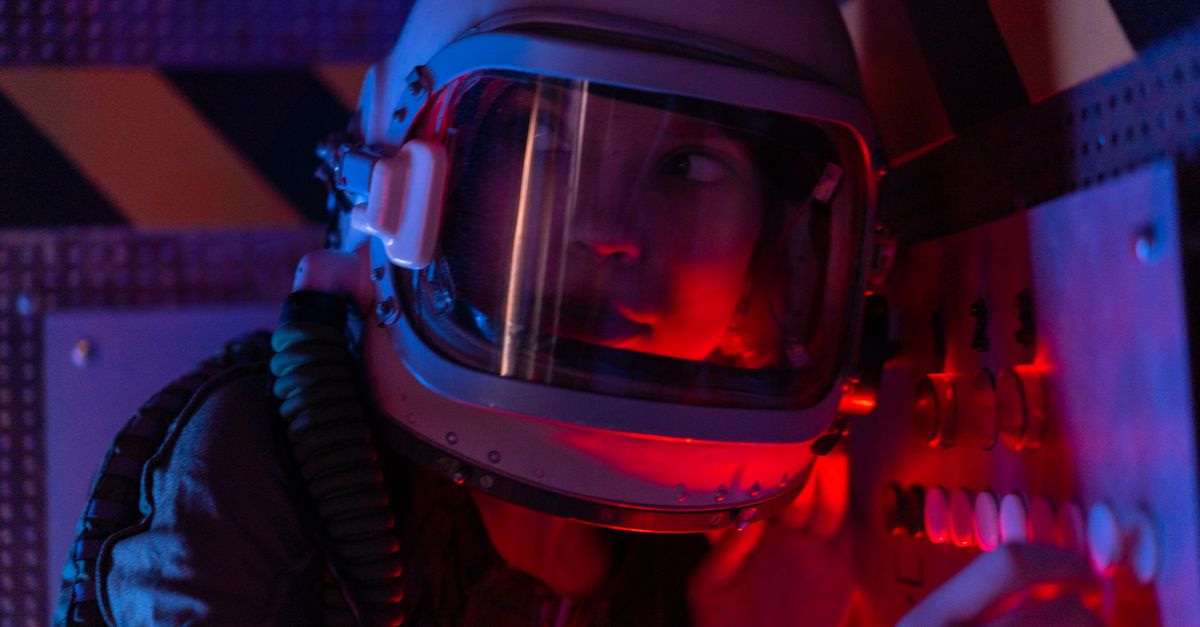 A woman astronaut wearing a spacesuit in a control room lit by red lights, looks focused.