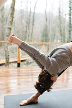 Woman performs yoga stretch outdoors on a wooden deck surrounded by trees.